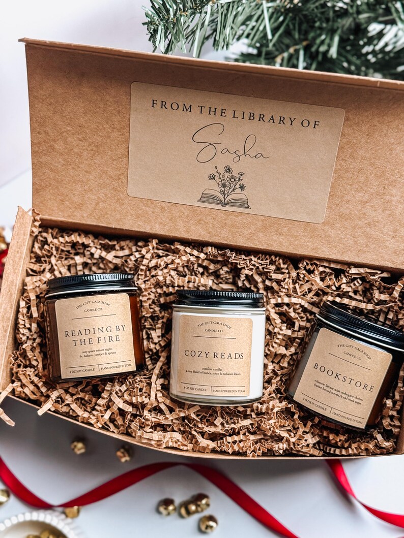 May include: A gift box filled with brown shredded paper and three candles. The candles are labeled "Reading by the Fire", "Cozy Reads", and "Bookstore". The box has a label that says "From the Library of Sasha".
