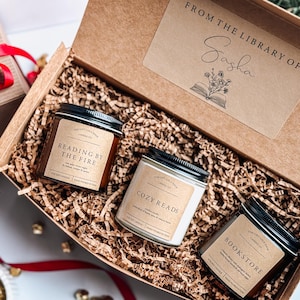 May include: A gift box filled with brown shredded paper and three candles. The candles are labeled "Reading by the Fire", "Cozy Reads", and "Bookstore". The box has a label that says "From the Library of Sasha".