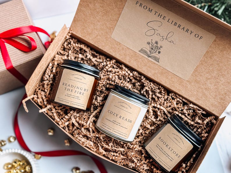 May include: A gift box filled with brown shredded paper and three candles. The candles are labeled "Reading by the Fire", "Cozy Reads", and "Bookstore". The box has a label that says "From the Library of Sasha".