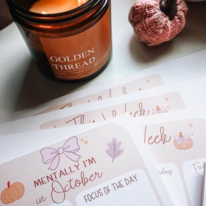 Pu&ograve; includere: Un barattolo di candela in vetro marrone con un'etichetta che riporta la scritta GOLDEN THREAD, una piccola zucca lavorata a maglia rosa e diversi planner di carta impilati con il testo MENTALLY I'M in October, TO-DO e FOCUS OF THE DAY con fiocchi.