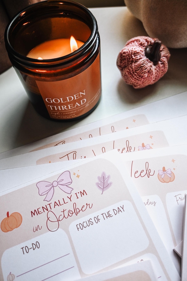 Pu&ograve; includere: Un barattolo di candela in vetro marrone con un'etichetta che riporta la scritta GOLDEN THREAD, una piccola zucca lavorata a maglia rosa e diversi planner di carta impilati con il testo MENTALLY I'M in October, TO-DO e FOCUS OF THE DAY con fiocchi.