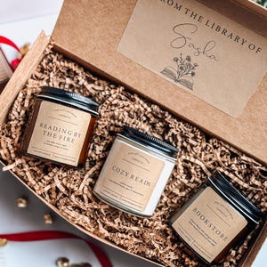 May include: A gift box filled with brown paper shred and three candles. The candles are labeled "Reading by the Fire", "Cozy Reads", and "Bookstore". The box has a label that says "From the Library of Sasha" with a drawing of an open book and flowers.