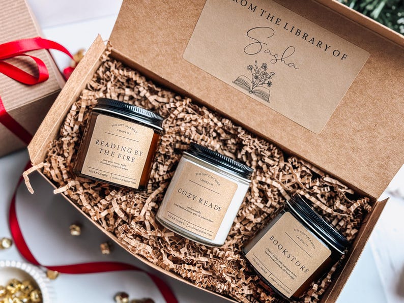 May include: A gift box filled with brown paper shred and three candles. The candles are labeled "Reading by the Fire", "Cozy Reads", and "Bookstore". The box has a label that says "From the Library of Sasha" with a drawing of an open book and flowers.
