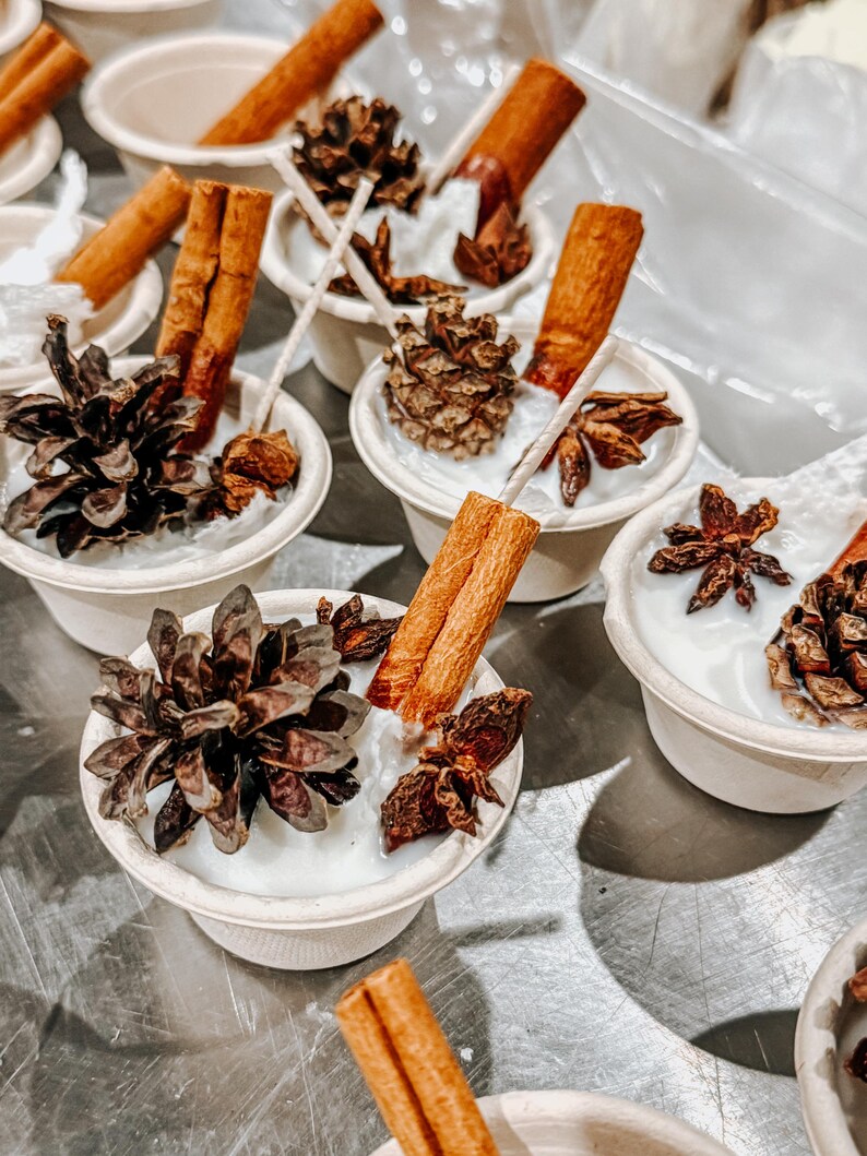 K&ouml;nnte beinhalten: Mehrere kleine, wei&szlig;e Kerzen in Pappbechern sind mit Zimtstangen, Tannenzapfen und Sternanis verziert. Die Kerzen haben eine rustikale, nat&uuml;rliche &Auml;sthetik, ideal f&uuml;r Heimdekoration oder Geschenke. Die Anordnung befindet sich auf einer metallischen Oberfl&auml;che.