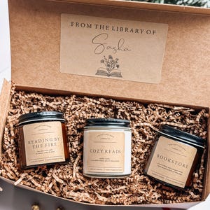 May include: A brown box with a label that says "From the Library of Sasha" and contains three candles. The candles are labeled "Reading by the Fire", "Cozy Reads", and "Bookstore".