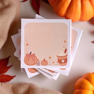 May include: A stack of square paper memo sheets featuring a light peach background with a white border. The bottom edge displays illustrations of a pumpkin with a bow, a small candle, a smaller pumpkin, and a cup of coffee with whipped cream.
