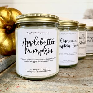 May include: Four glass candle jars with gold lids and white labels. The labels have handwritten text in black. The candles are labeled "Applebutter Pumpkin", "Cinnamon Pumpkin Cream", "Vanilla Strudel", and "Pumpkin Spice Latte". The candles are arranged in a row on a wooden surface.