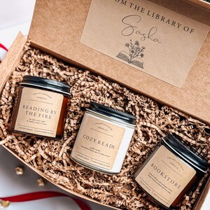 May include: A gift box filled with brown shredded paper and three soy candles. The candles are labeled "Reading by the Fire", "Cozy Reads", and "Bookstore". The box has a label that says "From the Library of Sasha" with a book and flowers illustration.