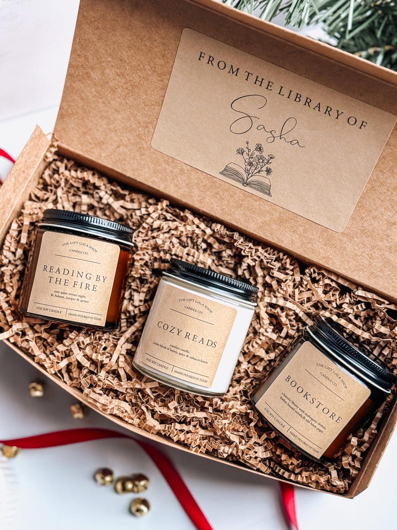 May include: A gift box filled with brown shredded paper and three soy candles. The candles are labeled "Reading by the Fire", "Cozy Reads", and "Bookstore". The box has a label that says "From the Library of Sasha" with a book and flowers illustration.