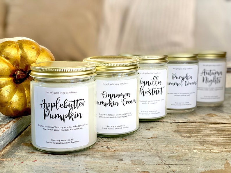 May include: Five glass jars with gold lids containing soy wax candles. The labels are white with black text and list the scents: Applebutter Pumpkin, Cinnamon Pumpkin Cream, Vanilla Chestnut, Pumpkin Caramel Dreams, and Autumn Nights. Each jar is 8 ounces and hand-poured in small batches.