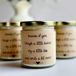 May include: A set of four glass candle jars with gold lids and brown paper labels. The label on the front jar reads "Because of you. I laugh a little harder, cry a little less, and smile a lot more!"