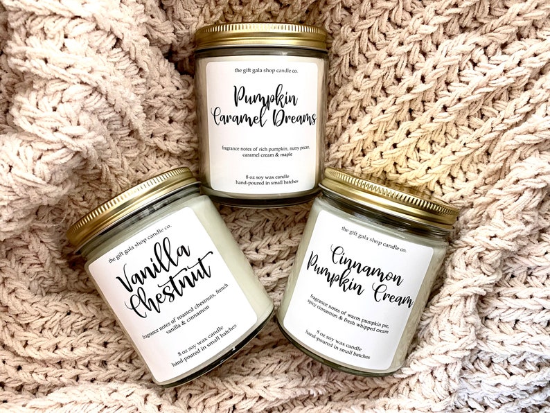 May include: Three 8-ounce soy wax candles in glass jars with gold lids. The candles are labeled "Pumpkin Caramel Dreams", "Cinnamon Pumpkin Cream", and "Vanilla Chestnut". Each label includes fragrance notes and the text "hand-poured in small batches".