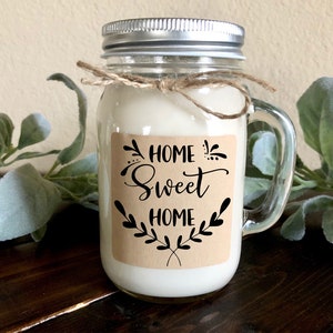 May include: A glass jar candle with a brown paper label that says "Home Sweet Home" with a decorative floral design. The candle has a silver lid and a clear glass handle.