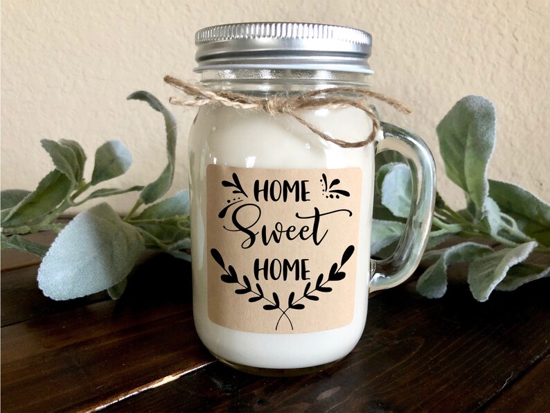 May include: A glass jar candle with a brown paper label that says "Home Sweet Home" with a decorative floral design. The candle has a silver lid and a clear glass handle.
