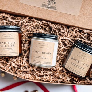 May include: Three candles in a brown box with shredded paper. The candles are labeled "Reading by the Fire", "Cozy Reads", and "Bookstore".