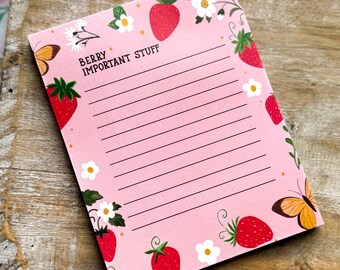 May include: A pink notepad with the text "BERRY IMPORTANT STUFF" and lined pages. The notepad is decorated with illustrations of strawberries, white flowers, and butterflies. The notepad is on a wooden tray.