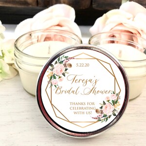 Pode incluir: Uma vela branca em um frasco de vidro transparente com um r&oacute;tulo com padr&atilde;o floral dourado que diz "Teresa's Bridal Shower 5.22.20 Thanks for celebrating with us!"