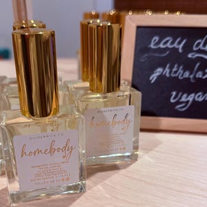 May include: Clear glass perfume bottles with gold-colored caps are arranged on a light-colored surface. Each bottle has a label with the word "homebody" in gold script. A small chalkboard displays the words "eau de parfum, phthalate free, vegan."