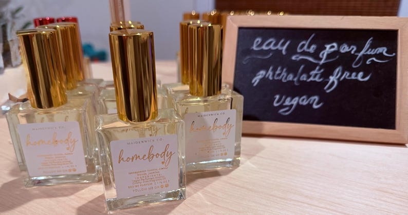 May include: Clear glass perfume bottles with gold-colored caps are arranged on a light-colored surface. Each bottle has a label with the word "homebody" in gold script. A small chalkboard displays the words "eau de parfum, phthalate free, vegan."