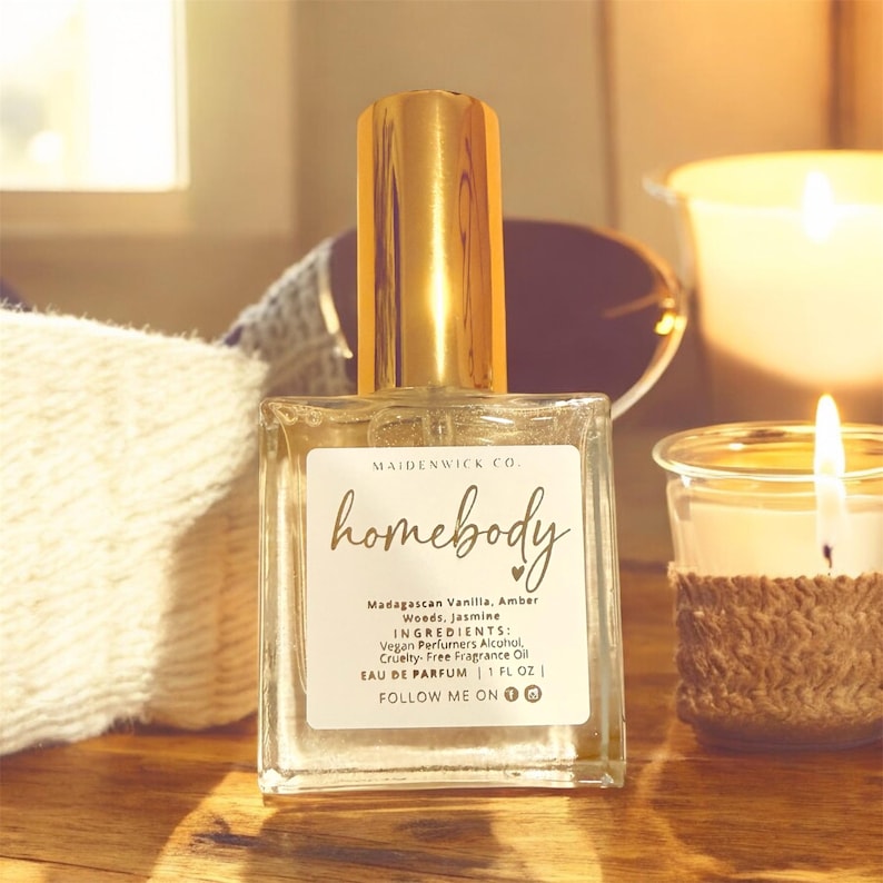 May include: A clear glass perfume bottle with a gold cap, labeled "homebody." The label lists ingredients and the text "EAU DE PARFUM | 1 FL OZ." The bottle sits on a wooden surface near a lit candle and a white cloth.