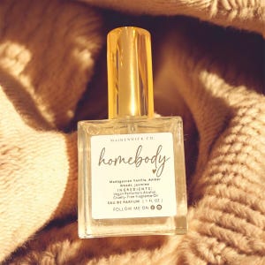 May include: A clear glass perfume bottle with a gold cap, labeled "homebody" with ingredients listed. The bottle is set against a soft, textured, beige background. The label includes the text "MAIDENWICK CO."