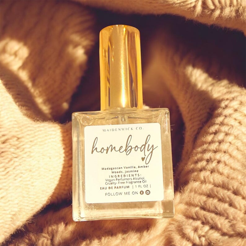 May include: A clear glass perfume bottle with a gold cap, labeled "homebody" with ingredients listed. The bottle is set against a soft, textured, beige background. The label includes the text "MAIDENWICK CO."