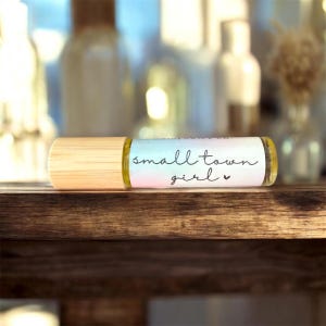 Könnte beinhalten: Eine transparente Glas-Rollerflasche für ätherische Öle mit hellem Holzdeckel. Auf dem Etikett steht "small town girl" in schwarzer Schrift. Die Flasche steht auf einer dunklen Holzoberfläche, mit verschwommenen Flaschen im Hintergrund.