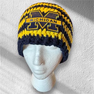 May include: A handmade crochet beanie in navy blue and yellow stripes. The beanie features a patch with the word "MICHIGAN" and the letter "M" in navy blue and yellow. The hat is displayed on a white mannequin head.