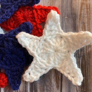 May include: Three crocheted stars in red, white, and blue, reminiscent of the American flag. The stars are arranged in a pile on a wooden surface.