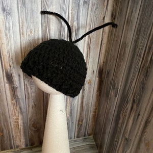 May include: Black crocheted ant hat with pipe cleaner antennae. The hat is displayed on a white styrofoam cone.