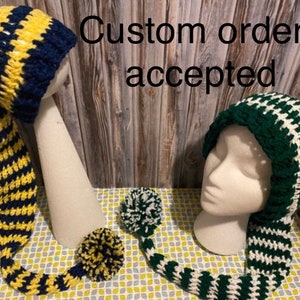 May include: Two crocheted hats with long tails. The hat on the left is blue and yellow striped. The hat on the right is green and white striped. The text "Custom orders accepted" is visible in the image.