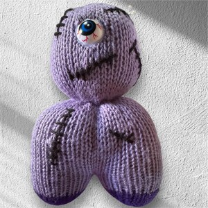 May include: A handmade, knitted purple doll with a single, large, googly eye. The doll features black stitched details and a stitched mouth. The doll's body is a lighter shade of purple, with darker purple accents at the bottom.