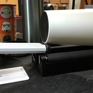 Op de afbeelding: Een witte plastic bekerhouder met een zwarte basis. De bekerhouder is ontworpen om een beker op zijn plaats te houden terwijl deze draait. De bekerhouder staat op een zwart oppervlak.