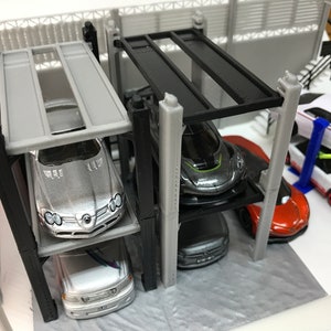 Peut inclure: Un garage &agrave; voitures miniature &agrave; plusieurs niveaux avec quatre &eacute;tages. Le garage est en plastique noir et gris et poss&egrave;de une structure en m&eacute;tal noir. Quatre voitures miniatures sont gar&eacute;es sur les diff&eacute;rents niveaux, dont une Mercedes-Benz argent&eacute;e, une voiture de sport grise, une voiture noire et une voiture orange.