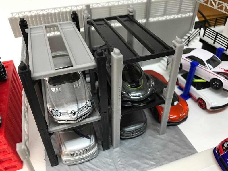Peut inclure: Un garage &agrave; voitures miniature &agrave; plusieurs niveaux avec quatre &eacute;tages. Le garage est en plastique noir et gris et poss&egrave;de une structure en m&eacute;tal noir. Quatre voitures miniatures sont gar&eacute;es sur les diff&eacute;rents niveaux, dont une Mercedes-Benz argent&eacute;e, une voiture de sport grise, une voiture noire et une voiture orange.