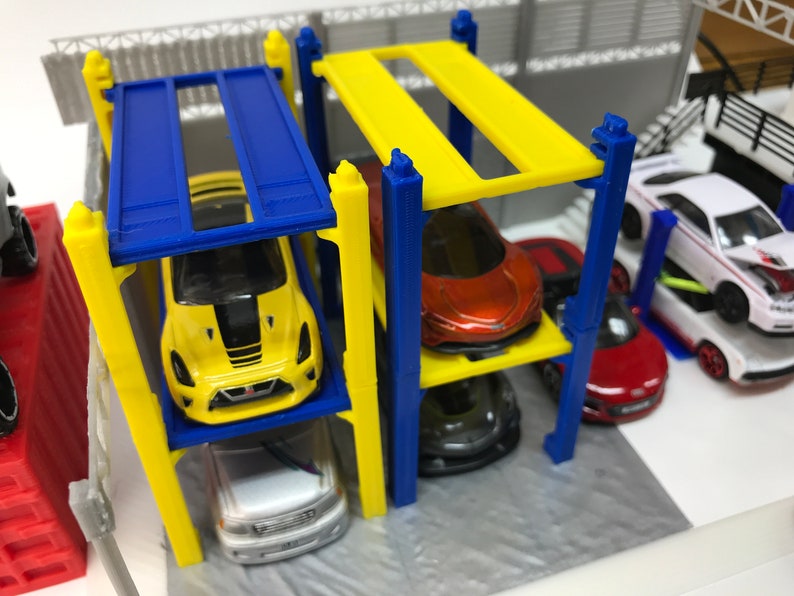 Peut inclure: Un garage de stationnement pour voitures miniatures jaune, bleu et blanc &agrave; quatre niveaux. Le garage est dot&eacute; de supports jaunes et bleus et de plateformes jaunes et bleues. Cinq voitures miniatures sont gar&eacute;es sur les plateformes, dont une jaune, une rouge, une blanche, une grise et une rouge.