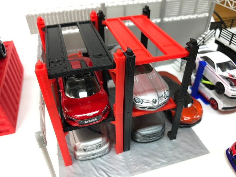Peut inclure: Un garage de stationnement pour voitures miniatures rouge, noir et argent avec quatre niveaux. Le garage est en plastique et poss&egrave;de un cadre en m&eacute;tal noir. Quatre voitures miniatures sont gar&eacute;es dans le garage, dont une voiture de sport rouge, une voiture de sport argent&eacute;e, une berline argent&eacute;e et une voiture de sport orange.