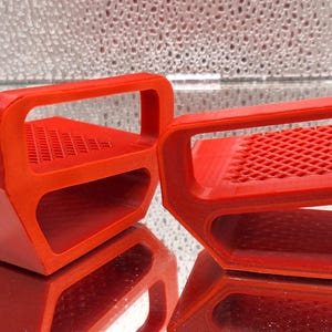 May include: Two bright red paint tray handles with a rectangular design and a handle. The handles have a grid-like pattern on the top surface. The objects are reflected on a shiny surface.