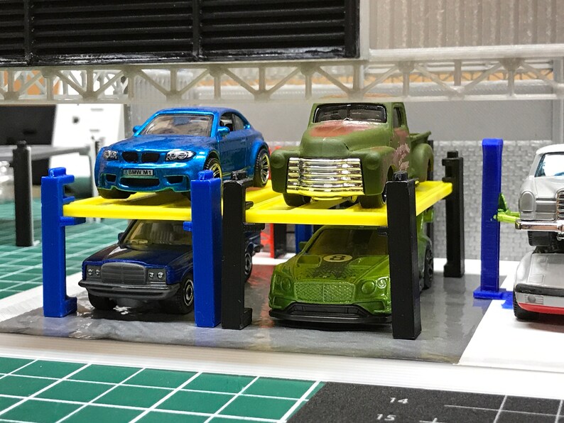 Peut inclure: Un &eacute;l&eacute;vateur de voiture jouet avec quatre voitures dessus. Les voitures sont une BMW M1 bleue, un pick-up vert, une Mercedes-Benz bleue et une Bentley verte.