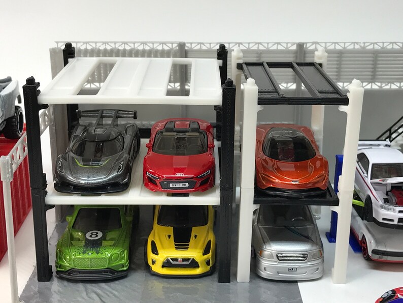 Peut inclure: Un garage &agrave; voitures &agrave; plusieurs niveaux blanc avec des poutres de soutien noires et un toit noir. Six voitures miniatures sont gar&eacute;es sur les diff&eacute;rents niveaux. Les voitures sont argent&eacute;es, rouges, orange, vertes, jaunes et argent&eacute;es.