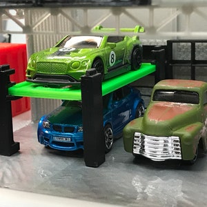 Peut inclure: Une voiture jouet verte avec un num&eacute;ro 8 blanc sur le c&ocirc;t&eacute; est sur une plateforme verte, une voiture jouet bleue est au niveau du sol, et un camion jouet vert est au niveau du sol.