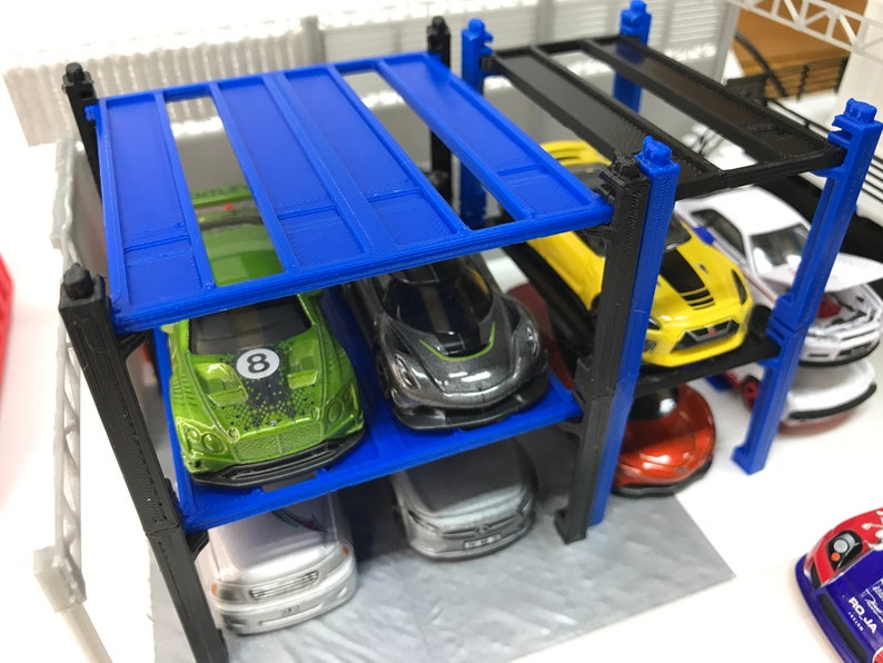Peut inclure: Un garage &agrave; voitures miniature &agrave; plusieurs niveaux avec des supports en m&eacute;tal bleu et noir. Le garage est rempli de voitures miniatures de diff&eacute;rentes couleurs, notamment vert, jaune, noir et blanc.