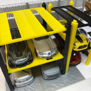 Peut inclure: Un garage &agrave; voitures &agrave; plusieurs niveaux jaune et noir avec quatre voitures miniatures gar&eacute;es &agrave; l'int&eacute;rieur. Les voitures sont une Mercedes-Benz argent&eacute;e, une voiture de sport noire et verte, une berline argent&eacute;e et une voiture de sport rouge.