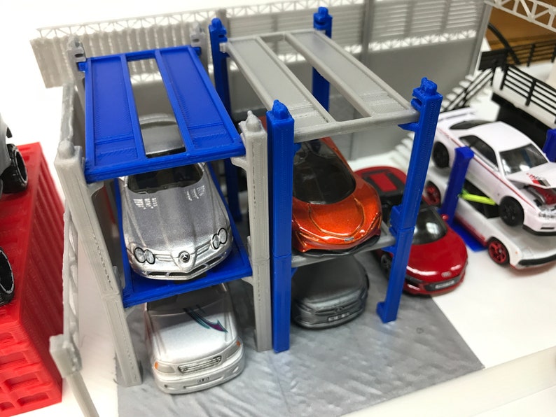 Peut inclure: Un jouet de garage &agrave; plusieurs niveaux avec quatre voitures miniatures. Le garage est en plastique gris avec des accents bleus. Les voitures sont argent&eacute;es, orange, rouge et blanche.