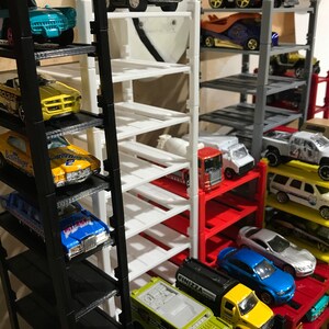 Peut inclure: Un garage &agrave; voitures miniature &agrave; plusieurs niveaux avec des &eacute;tag&egrave;res noires, blanches, rouges et grises. Le garage est rempli de voitures miniatures de diff&eacute;rentes couleurs et styles, dont un camion jaune avec l'inscription "FINIZZA" sur le c&ocirc;t&eacute;.