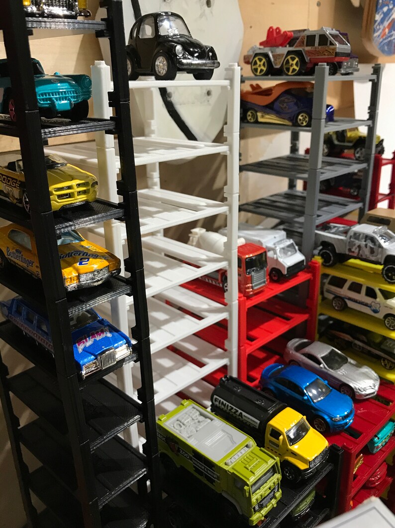 Peut inclure: Un garage &agrave; voitures miniature &agrave; plusieurs niveaux avec des &eacute;tag&egrave;res noires, blanches, rouges et grises. Le garage est rempli de voitures miniatures de diff&eacute;rentes couleurs et styles, dont un camion jaune avec l'inscription "FINIZZA" sur le c&ocirc;t&eacute;.