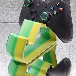 Peut inclure: Manette Xbox noire avec un support imprimé en 3D vert et jaune. La manette a des boutons colorés et des joysticks verts. Le support est une conception en couches avec un dégradé vert et jaune.