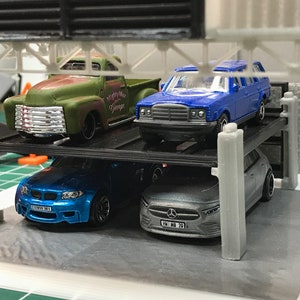Peut inclure: Un garage &agrave; voitures &agrave; plusieurs niveaux avec quatre voitures miniatures. Les voitures sont un pick-up vert, une berline bleue, une voiture de sport bleue et une berline argent&eacute;e. Le garage est en plastique noir avec des piliers gris.