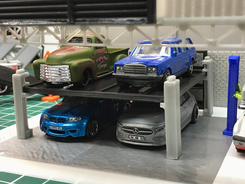 Peut inclure: Un garage &agrave; voitures &agrave; plusieurs niveaux avec quatre voitures miniatures. Les voitures sont un pick-up vert, une berline bleue, une voiture de sport bleue et une berline argent&eacute;e. Le garage est en plastique noir avec des piliers gris.