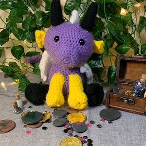 May include: Purple and yellow crocheted dragon plush toy sitting on a gray surface with gold and black coins scattered around it.